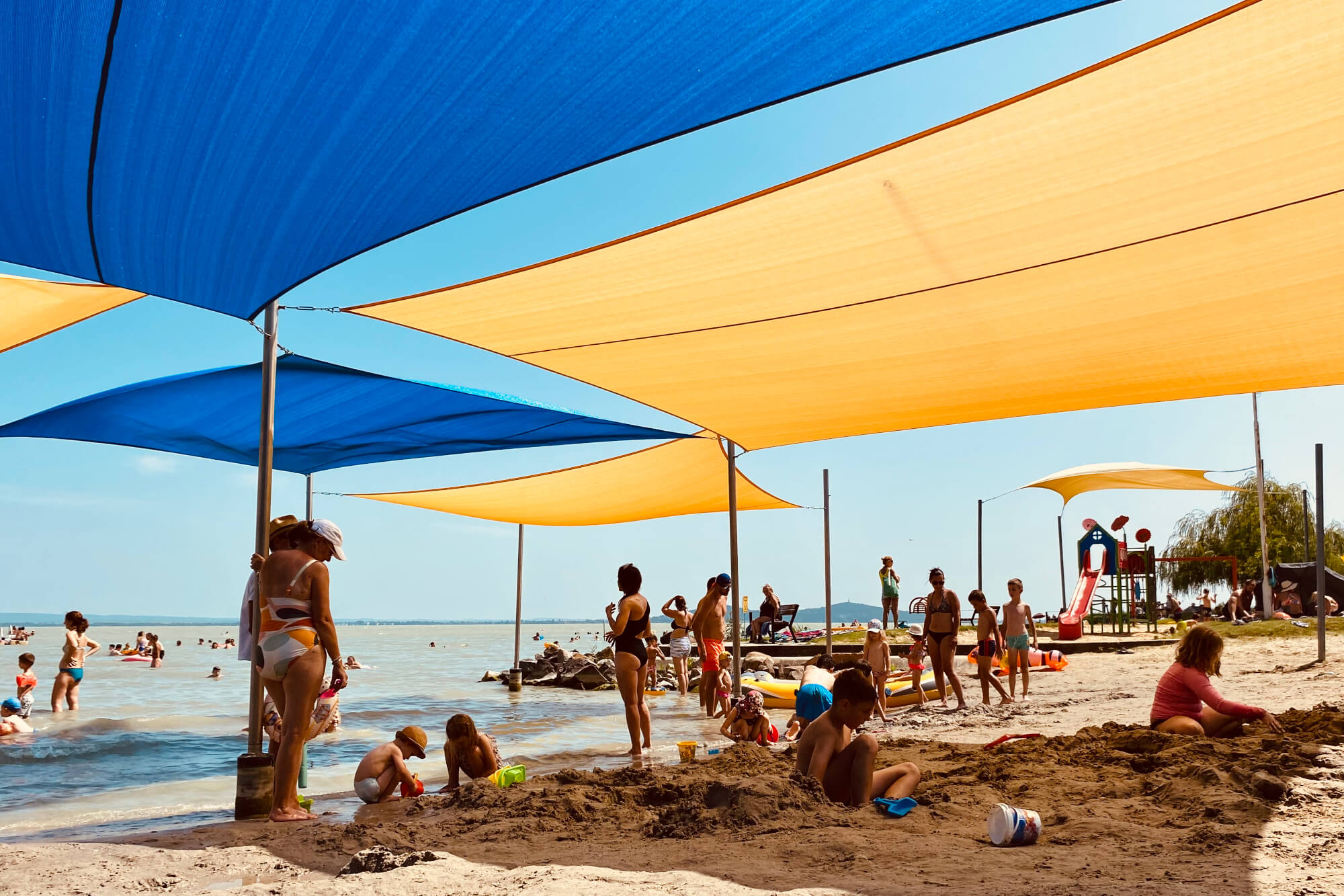 Napvitorla játszótér árnyékolás 5 Gyerekek és családok játszanak egy strandon, ahol színes napvitorlák biztosítanak árnyékot a part mentén